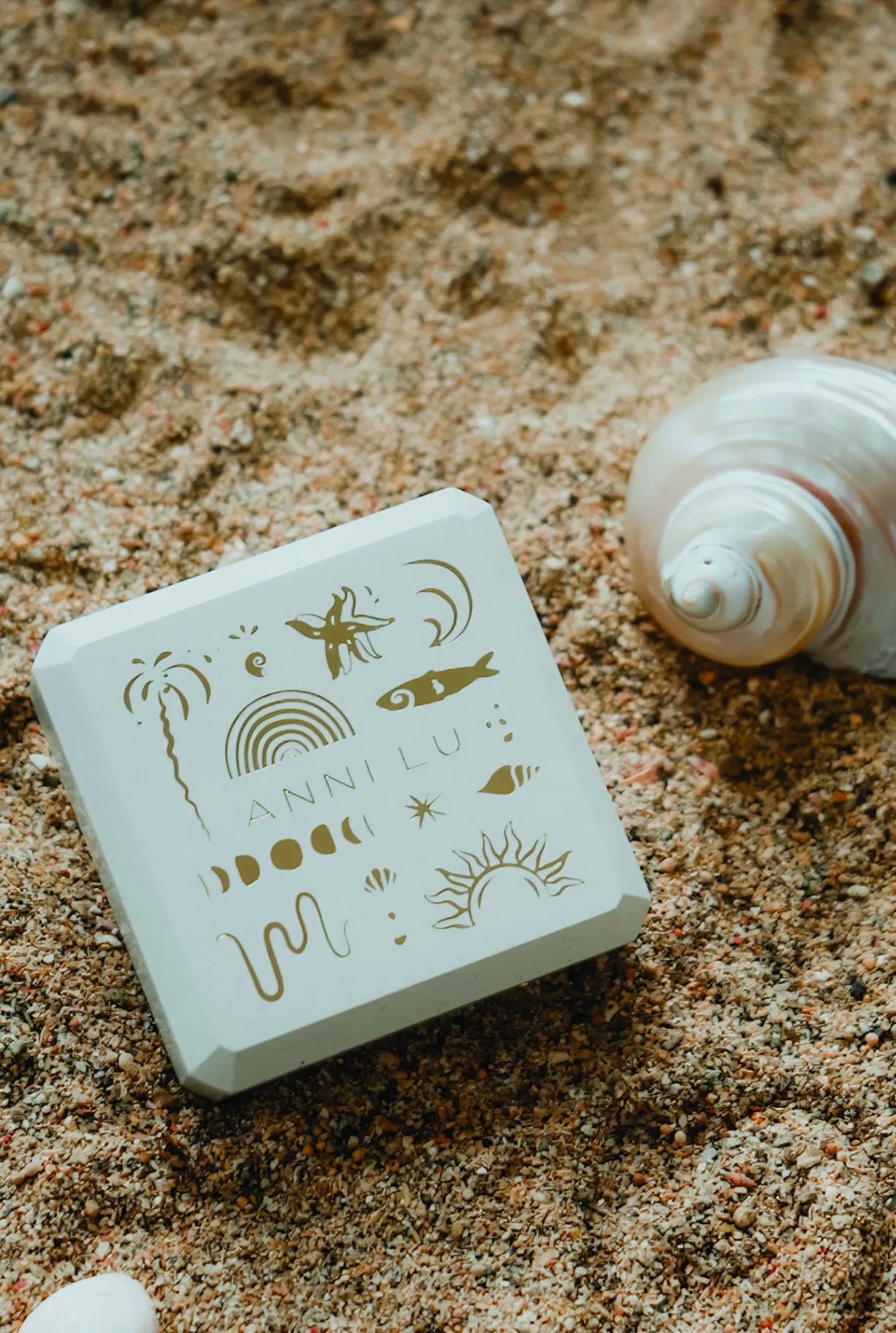Anni Lu jewellery box with gold foil illustrations resting on sand beside a seashell